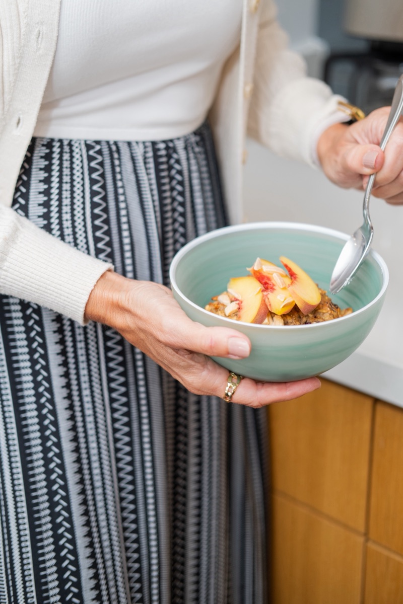 A woman in a cream cardigan holding a mint bowl of cookie crumble topped with fresh peach slices, spoon in hand